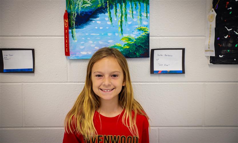 A girl wearing a red shirt smiles while standing against a wall